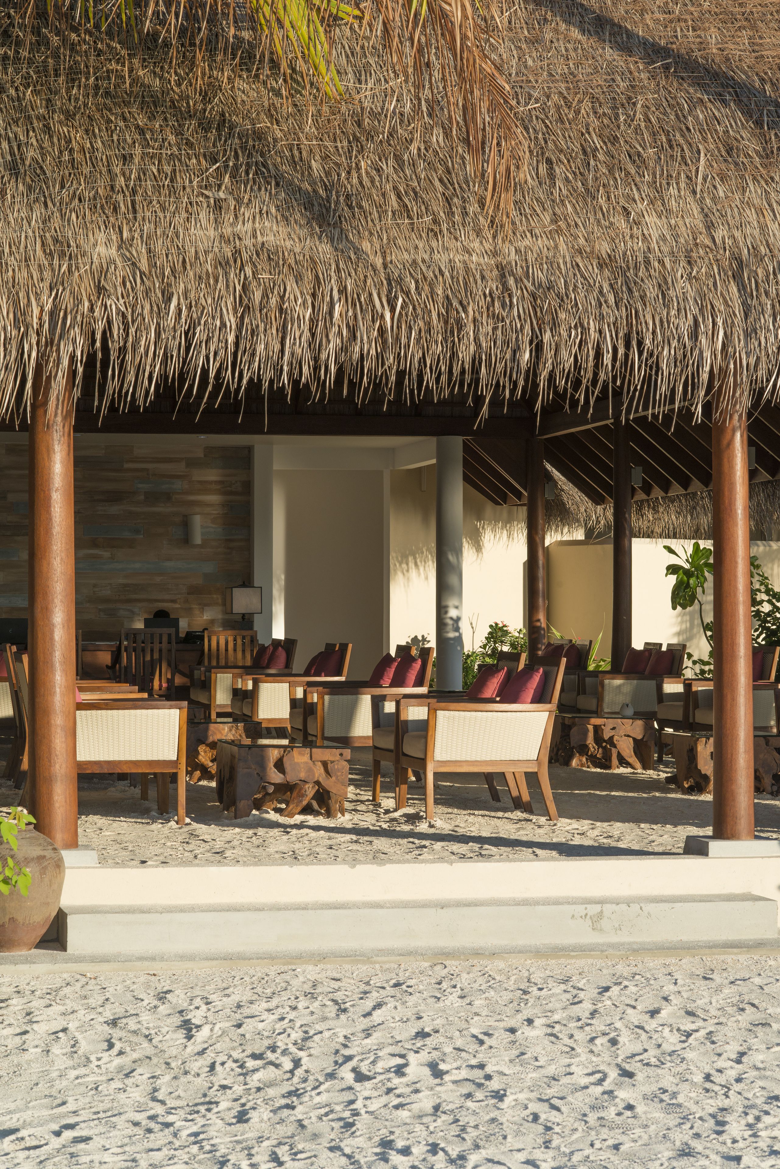 Un área de descanso junto a la playa con techos de paja, asientos y mesas de madera, rodeado de arena y palmeras.