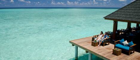 An overwater bungalows with wooden deck seating and lounge chairs, overlooking clear turquoise waters.