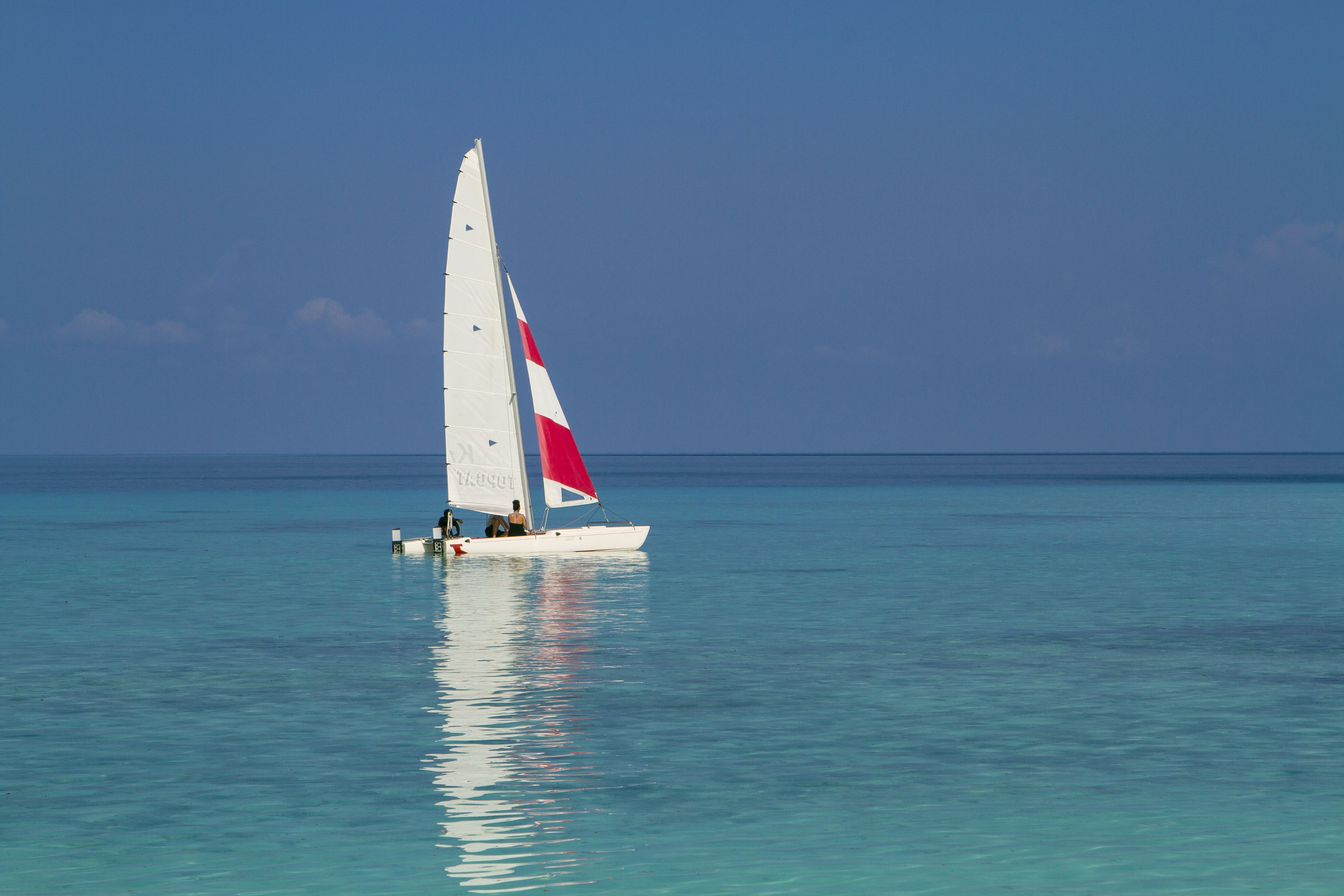 Un velero con vela roja y blanca navega sobre aguas turquesas tranquilas bajo un cielo despejado.