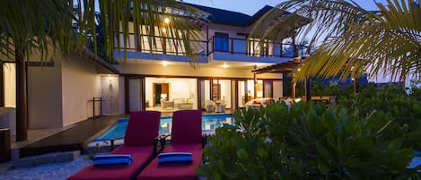Ein modernes, zweistöckiges Haus mit Swimmingpool, roten Liegestühlen und tropischen Pflanzen.