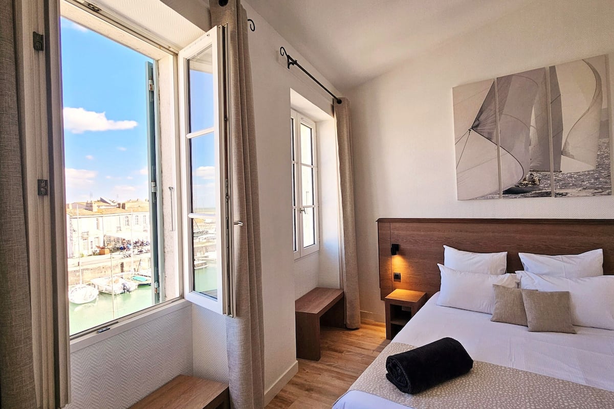 Una habitación de hotel con una cama, una mesita de noche, un ventanal con cortinas, un cuadro en la pared y vistas a edificios y agua.