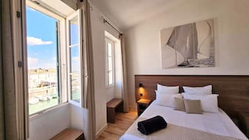 Una habitación de hotel con una cama, una mesita de noche, un ventanal con cortinas, un cuadro en la pared y vistas a edificios y agua.