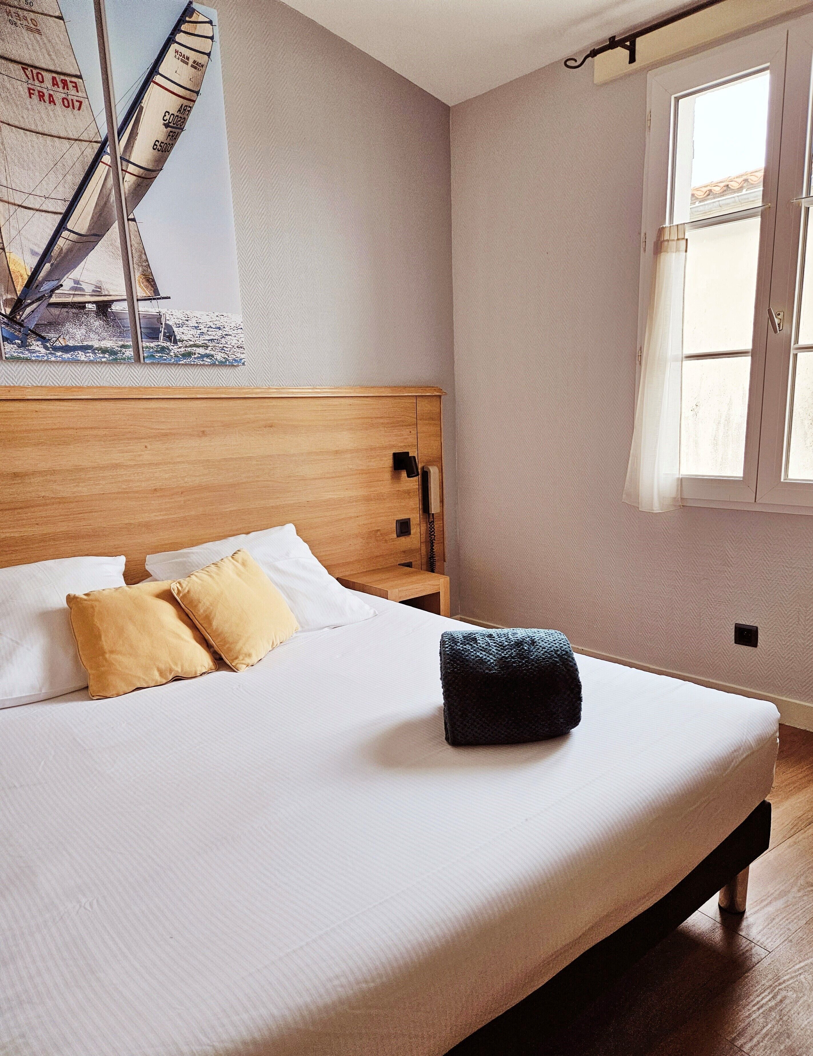 A bedroom with a wooden headboard, a bed with white and mustard pillows, a nightstand with a telephone, and a window with curtains.