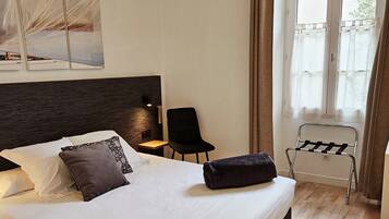 A neatly made bed with white linens and a dark headboard, a small bedside table with a lamp, a chair, and a window with curtains.