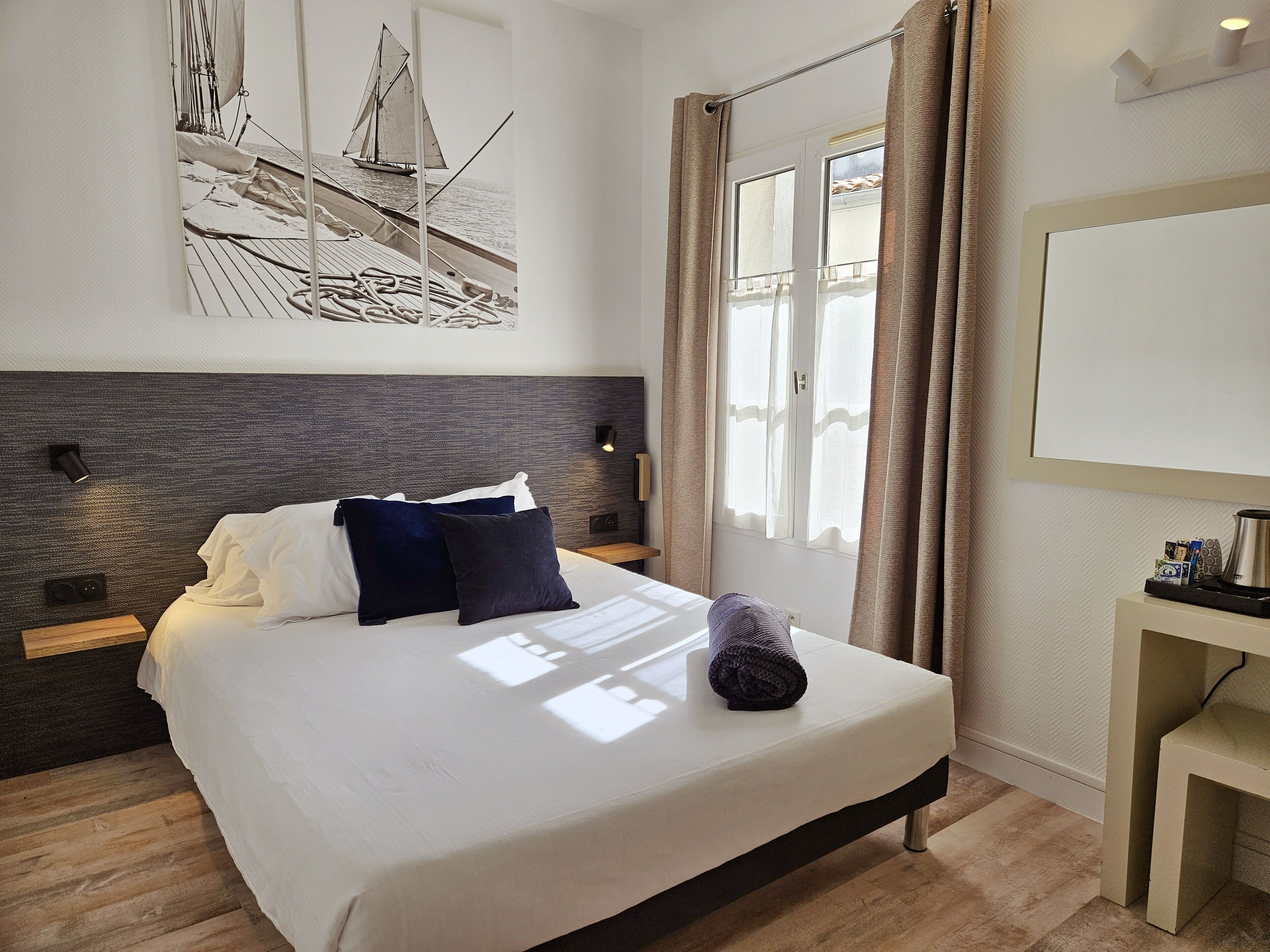Una habitación de hotel moderna con una cama grande, cabecera con diseño texturizado, una obra de arte colgada en la pared de un velero, una ventana con cortinas y una mesita de noche con lámpara.
