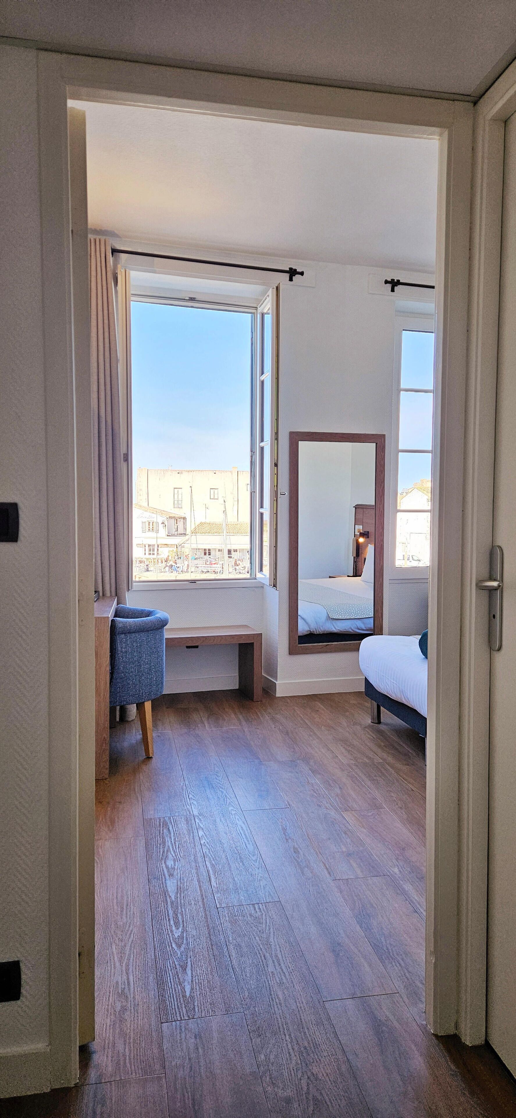Een kamer met een houten vloer, een bed met hoofdeinde, een bankje, een stoel en een groot raam met uitzicht op gebouwen.