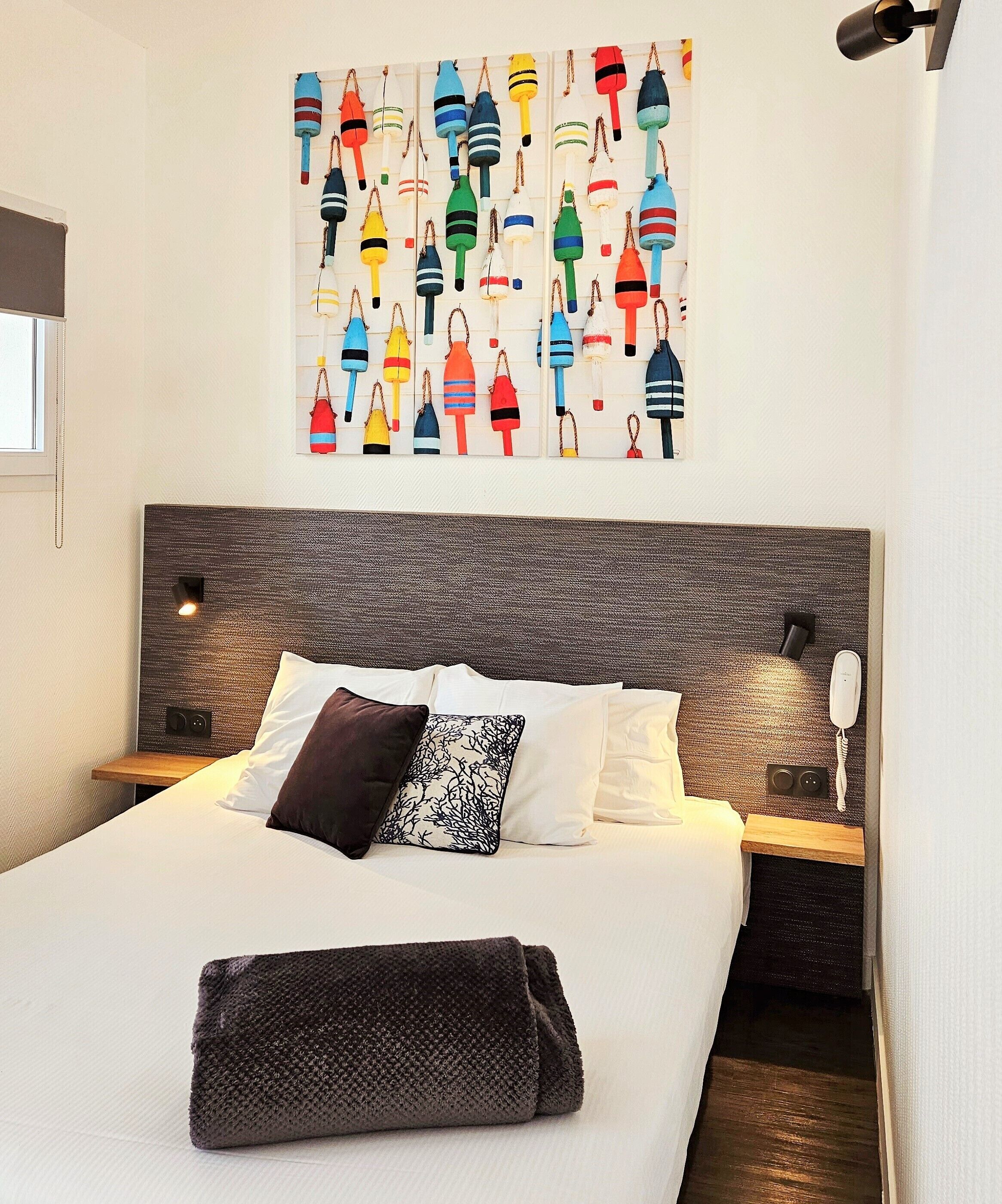 A modern hotel room with a large bed, a headboard with a textured pattern, a wall-mounted artwork of colorful buoys, and a bedside table with a lamp.