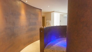 Uma escadaria em curva com piso de pedra e corrimão iluminado em azul, que leva a um espaço com parede branca e porta.