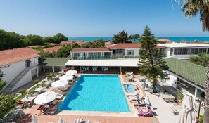 A rectangular swimming pool with lounge chairs and umbrellas, surrounded by buildings with red roofs and a clear view of the sea. - Clover Magic Nova Beach Hotel (Manavgat)