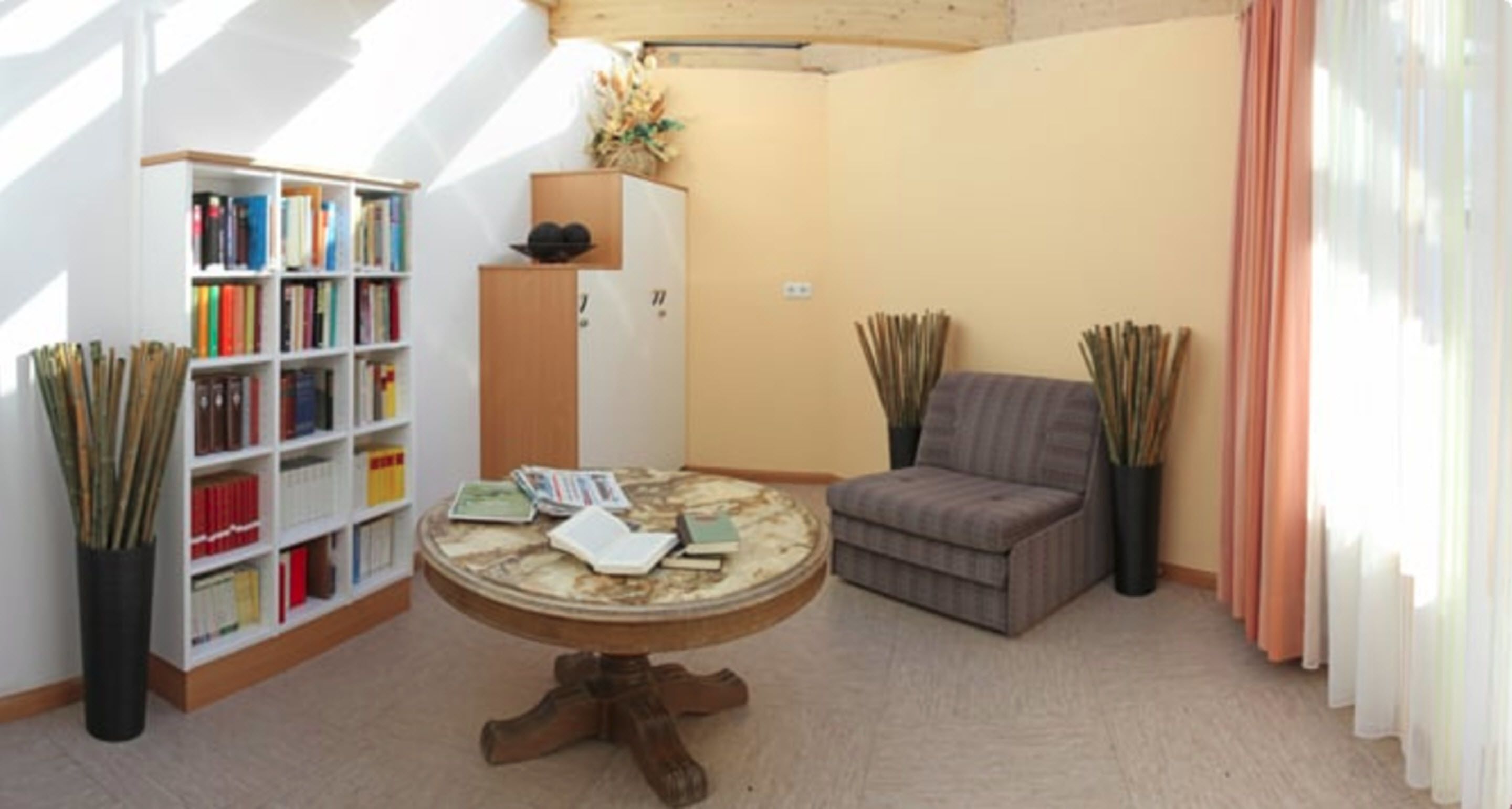 A room with a bookshelf, a round table with books, a grey sofa, and tall plants.