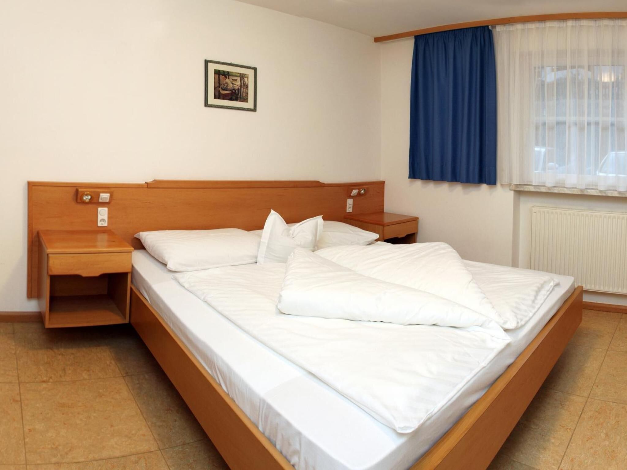 A single-bed room with a wooden headboard, two bedside tables, a small bedside drawer, a window with blue curtains, and a framed picture on the wall.