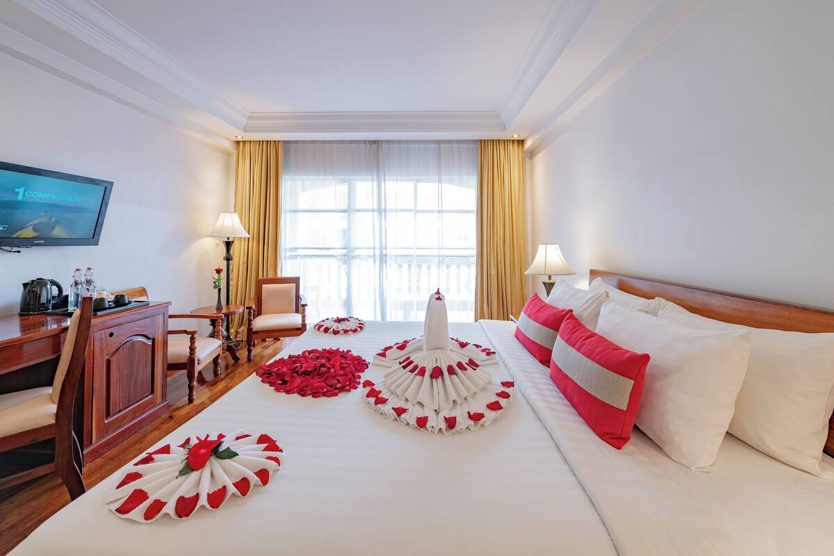 A hotel room with a neatly made bed, a television on the wall, a desk with a coffee maker, and a window with curtains.