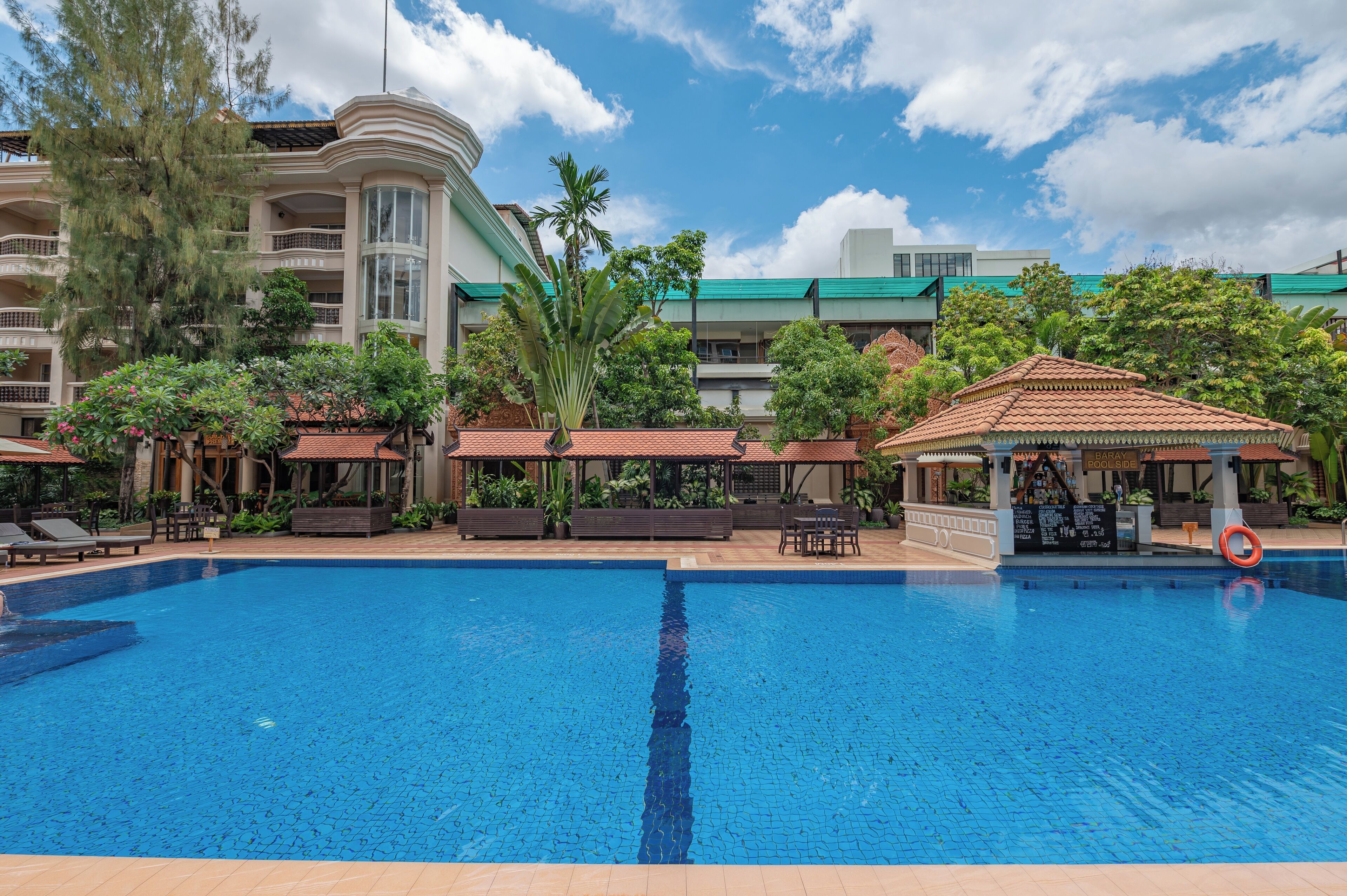 A swimming pool with lounge chairs and a bar area, surrounded by greenery and a multi-story building with balconies.