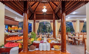 A buffet area with a variety of fruits and dishes, wooden pillars, and a view through large windows. - Somadevi Angkor Premium (Siem Reap)