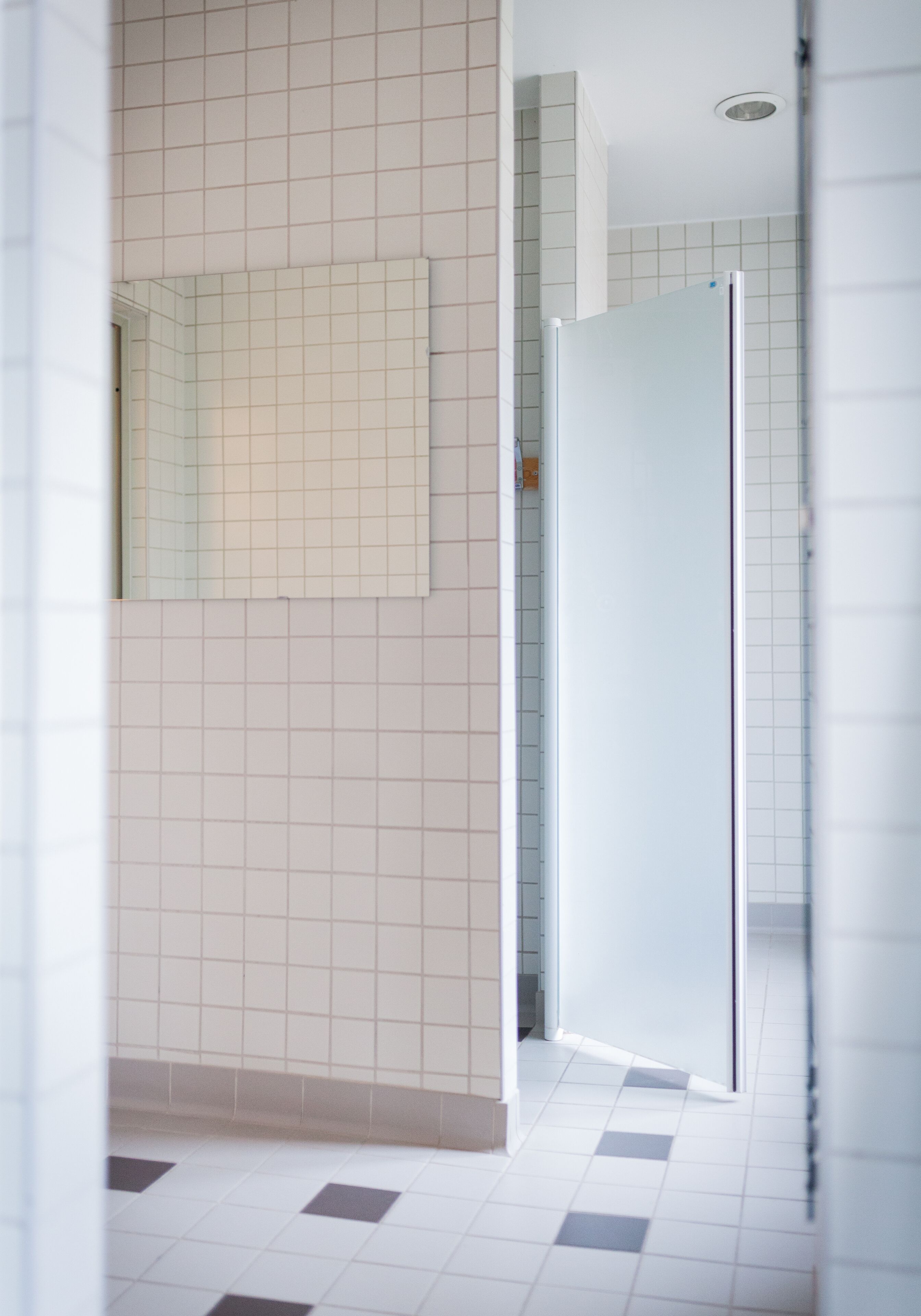 A bathroom with white tiled walls and floor, a mirror, and a door leading to another room.