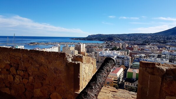 Un paisaje urbano costero con un muro de piedra histórico en primer plano, un puerto deportivo con barcos y una mezcla de edificios modernos y tradicionales.