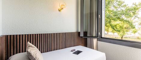 A neatly made bed with a pillow, a tablet, and glasses on it, a wall-mounted light, and a window with a view of trees.