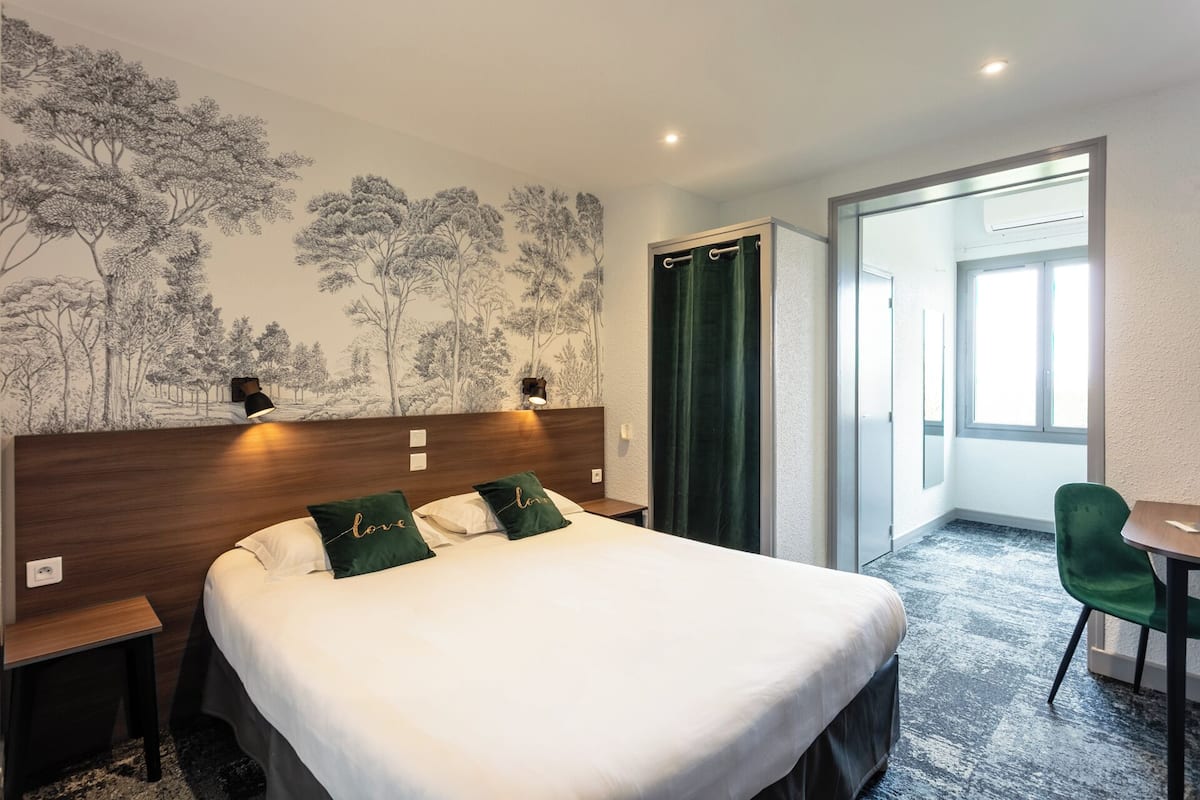 Habitación de hotel con una cama grande, un escritorio y una silla. Hay un mural en la pared con árboles y una puerta que da a otra habitación.