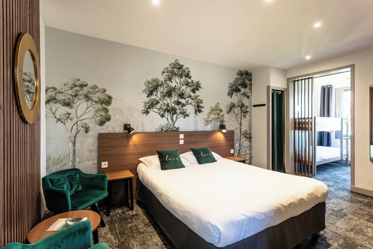 Habitación de hotel con una cama grande, cabecero de madera, un sillón verde, una mesita pequeña y una pared con un mural de un árbol.