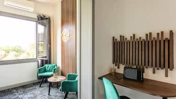 A hotel room with a large window, two green chairs, a small round table, a wooden wall panel, and a wooden shelf with a radio.