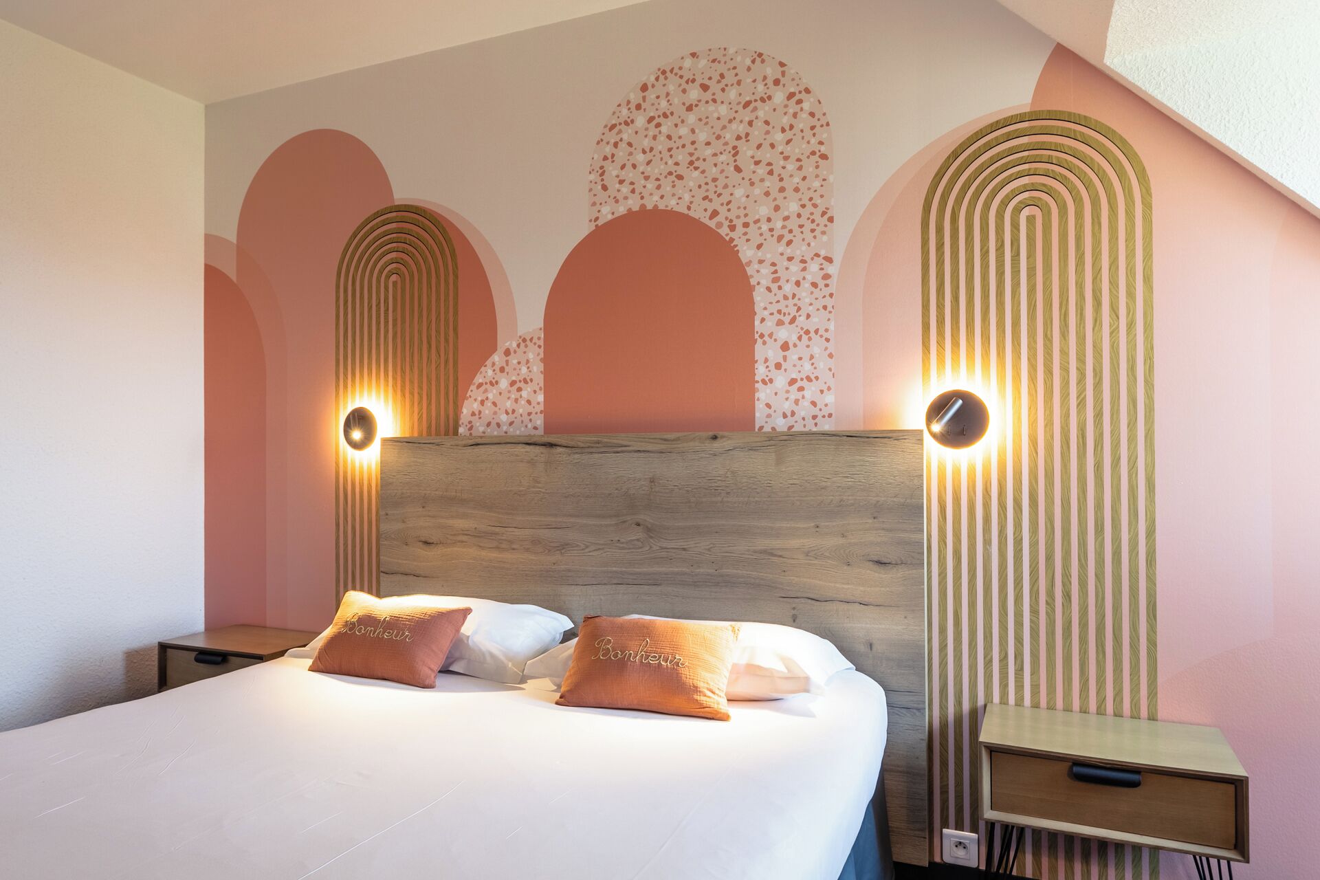 Un dormitorio con cabecera de madera, dos lámparas de noche, una mesita de noche y una cama con ropa blanca y dos almohadas naranjas.