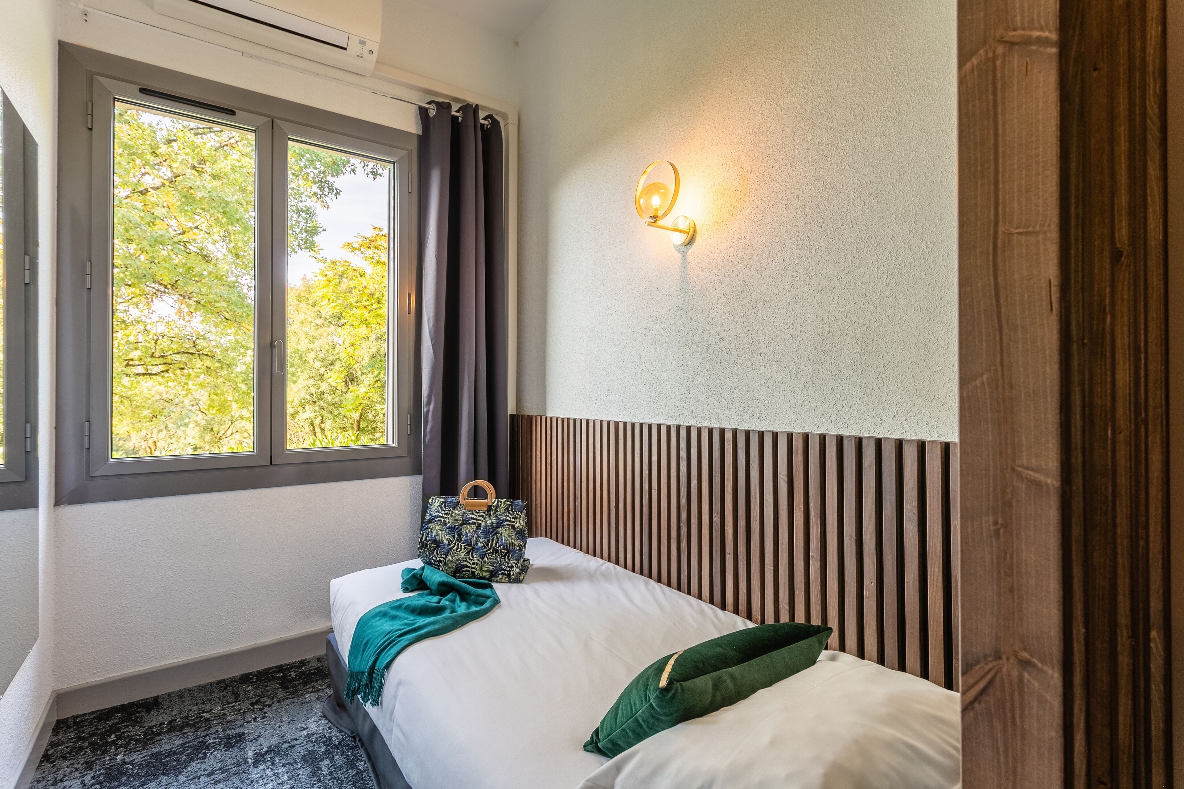 Een slaapkamer met een bed, een raam met gordijnen, een wandlamp en een houten hoofdbord.