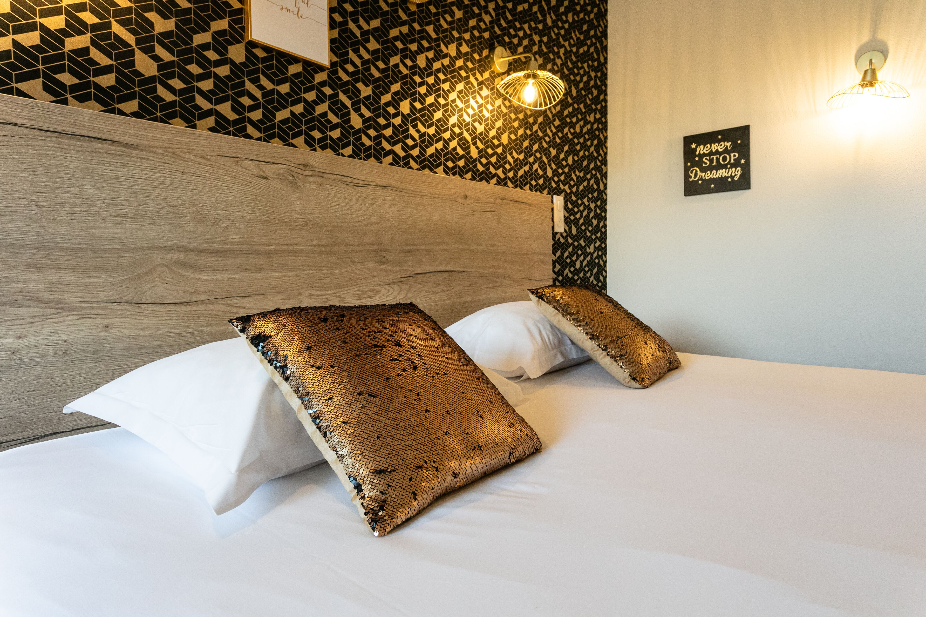 Un lit bien fait, avec une tête de lit en bois, un mur décoratif orné d’un motif géométrique et un panneau d’affichage affichant un message motivant.