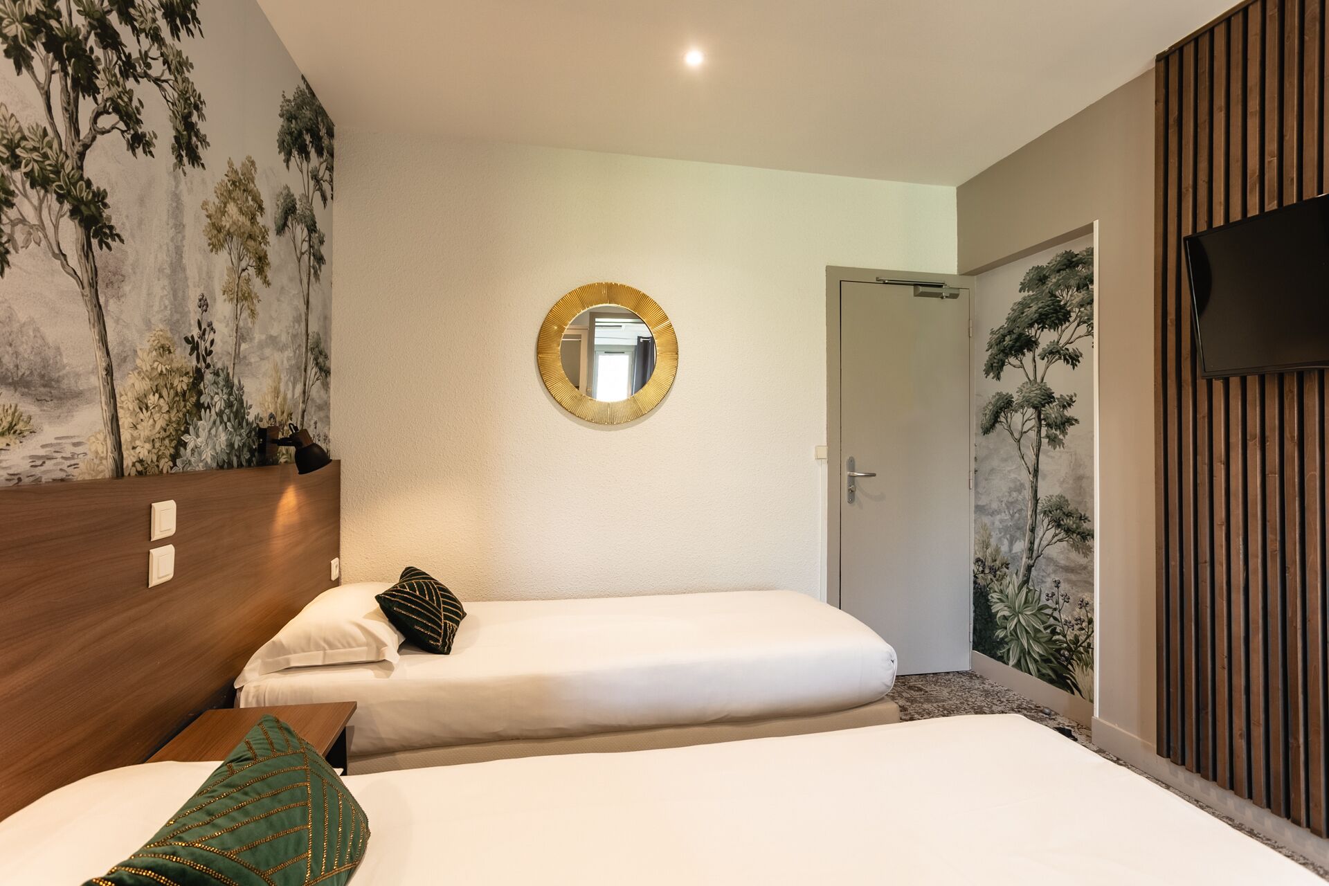 A hotel room with two beds, a round mirror, a TV, and a mural of trees on the wall.