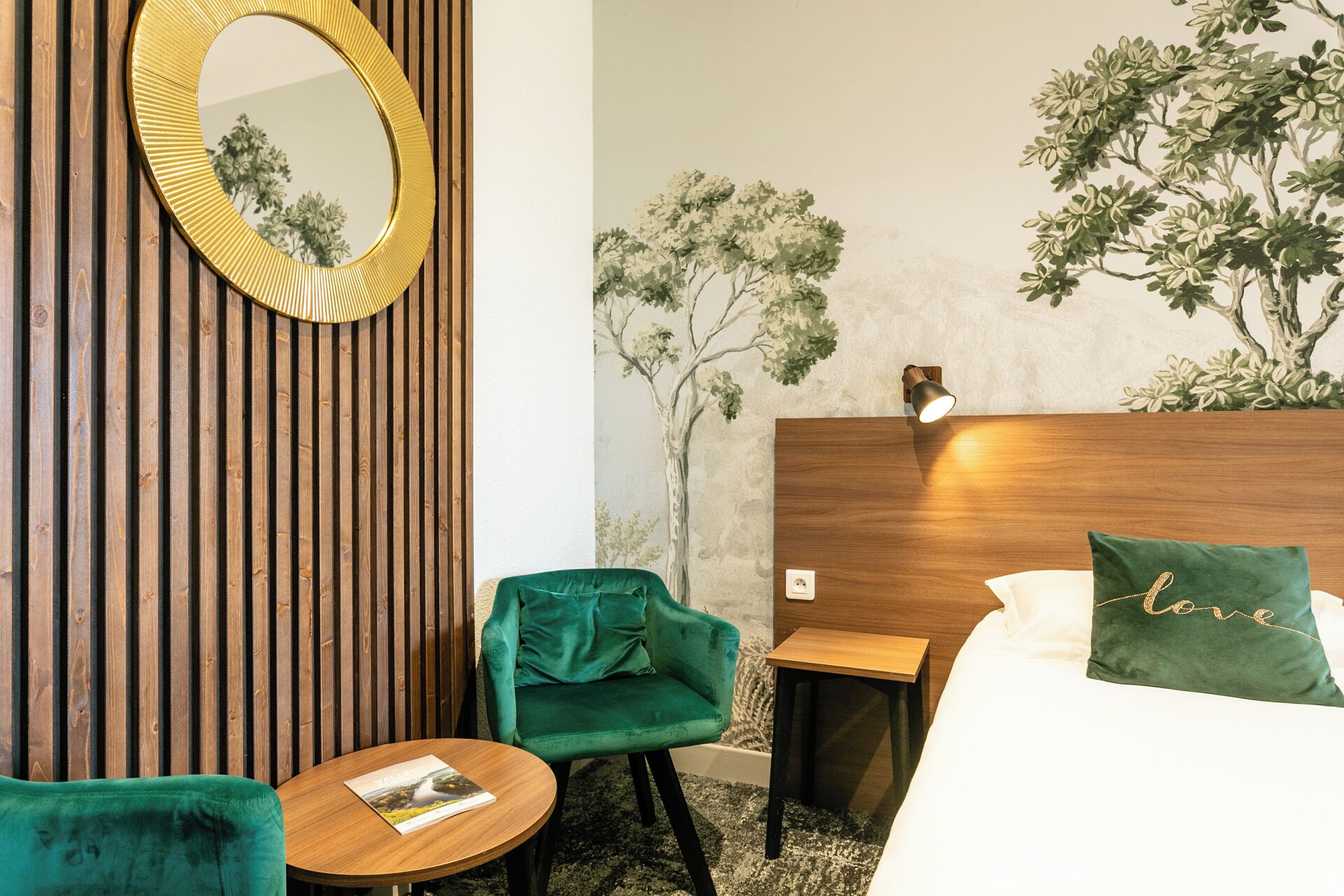 Une chambre d’hôtel avec un mur recouvert de lambris en bois, un miroir rond, une chaise verte, une petite table en bois, un lit avec des coussins verts et une fresque murale représentant des arbres.