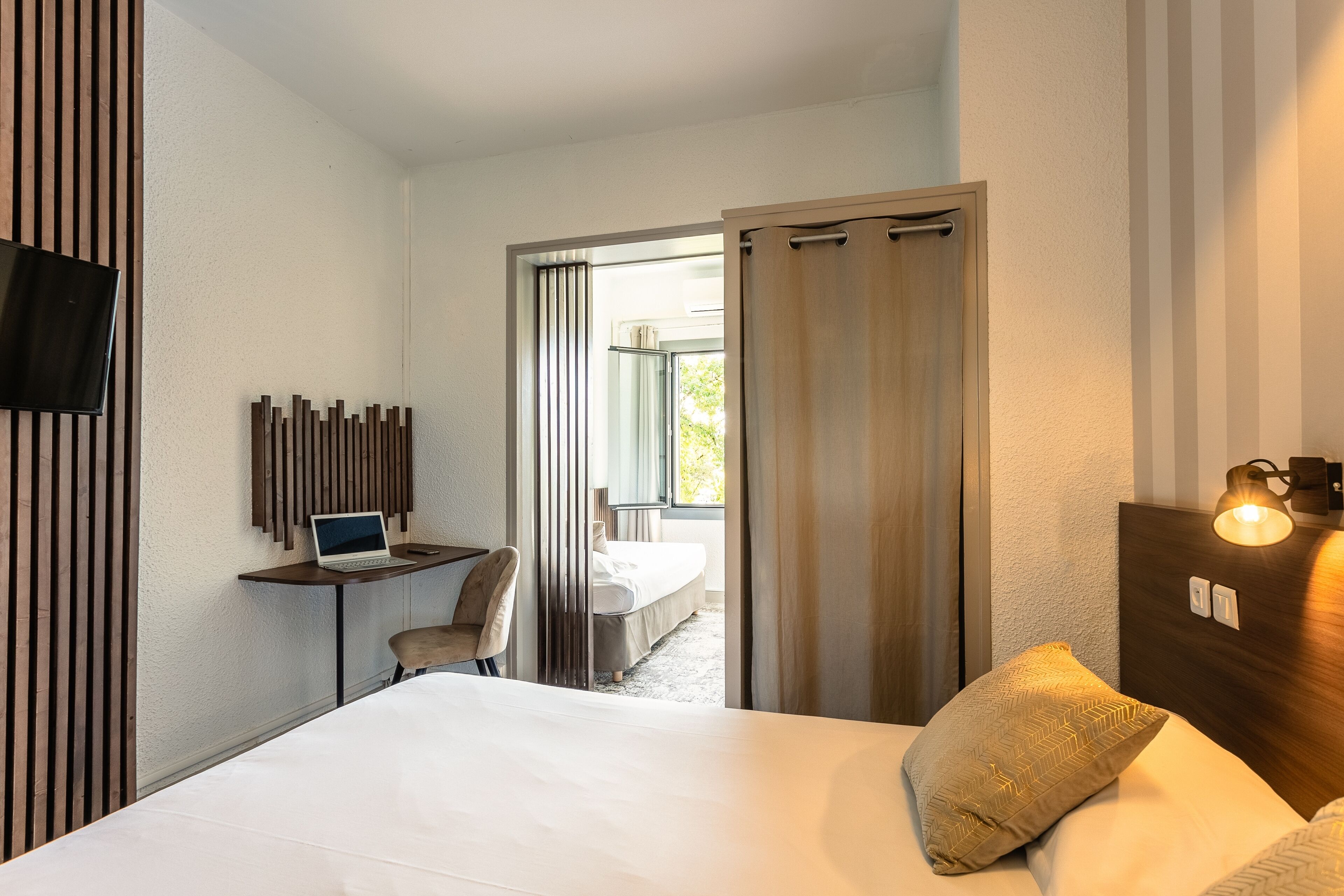 Una habitació d'hotel amb un llit, un escriptori amb un ordinador portàtil, una cadira, un televisor muntat a la paret i una finestra amb cortines.