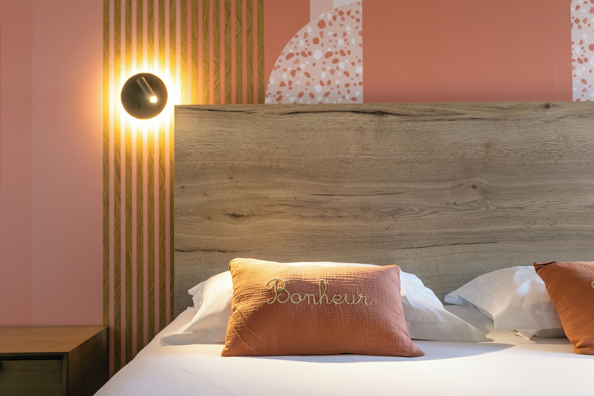 A neatly made bed with a wooden headboard, a single pillow with embroidered text, and a wall-mounted light fixture.