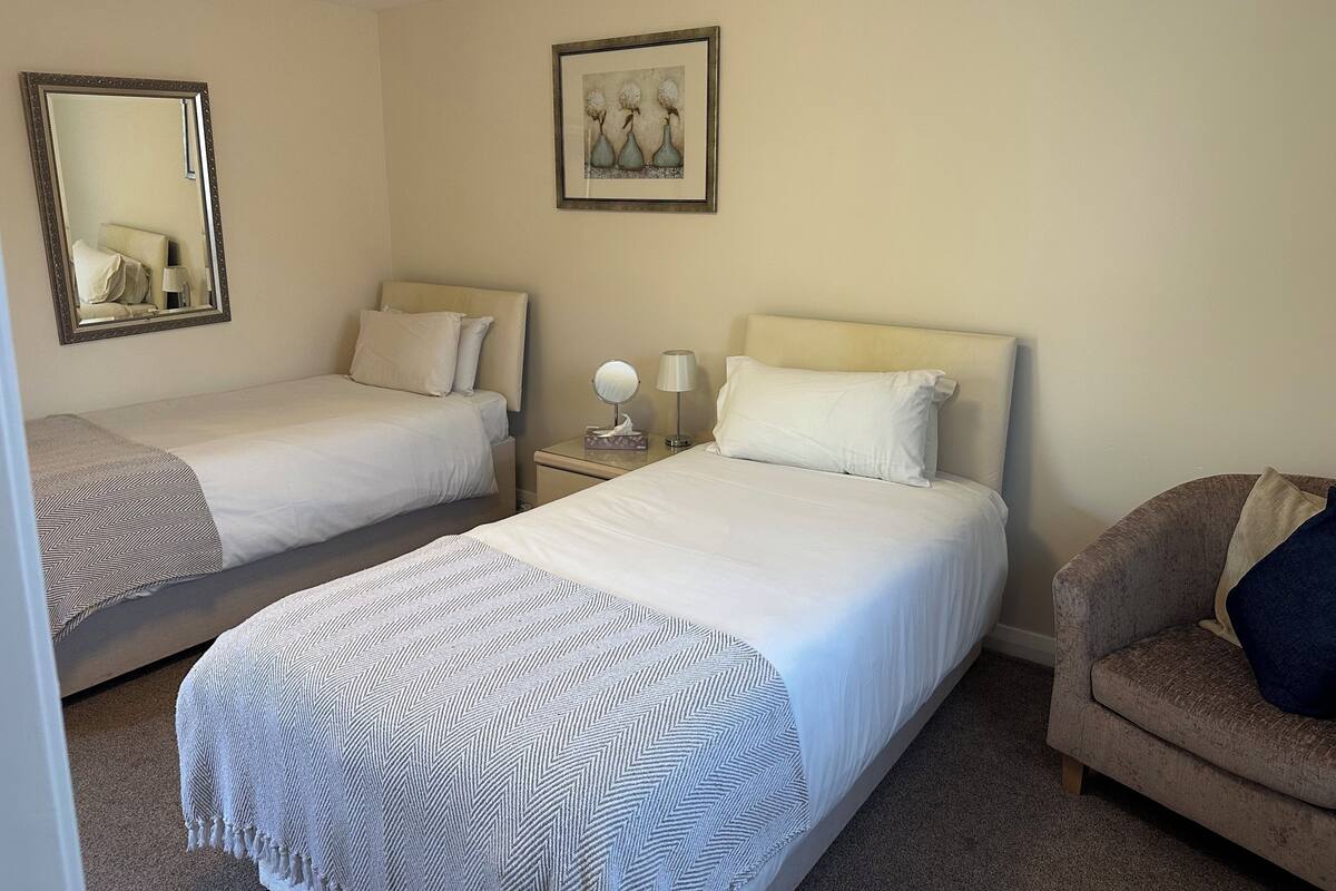 A hotel room with two single beds, a nightstand with a lamp, a chair, and a framed picture on the wall.