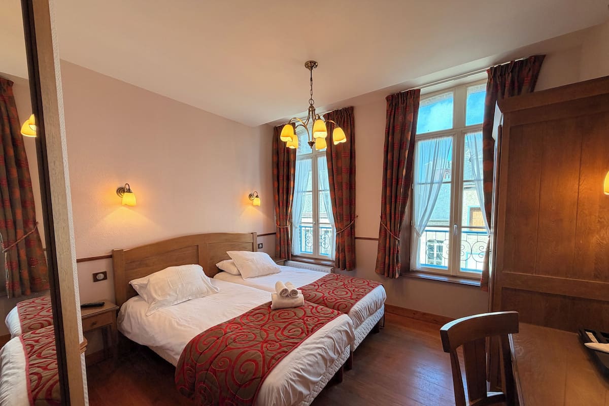 A hotel room with two beds, a wooden headboard, a dining table with a chair, and a window with curtains.
