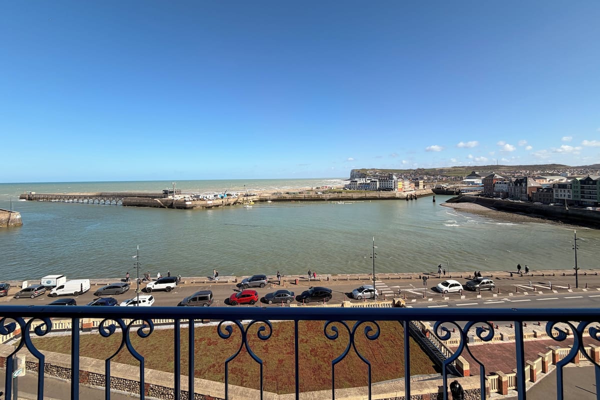 A coastal cityscape with a pier, waterfront parking, and buildings along the shore.
