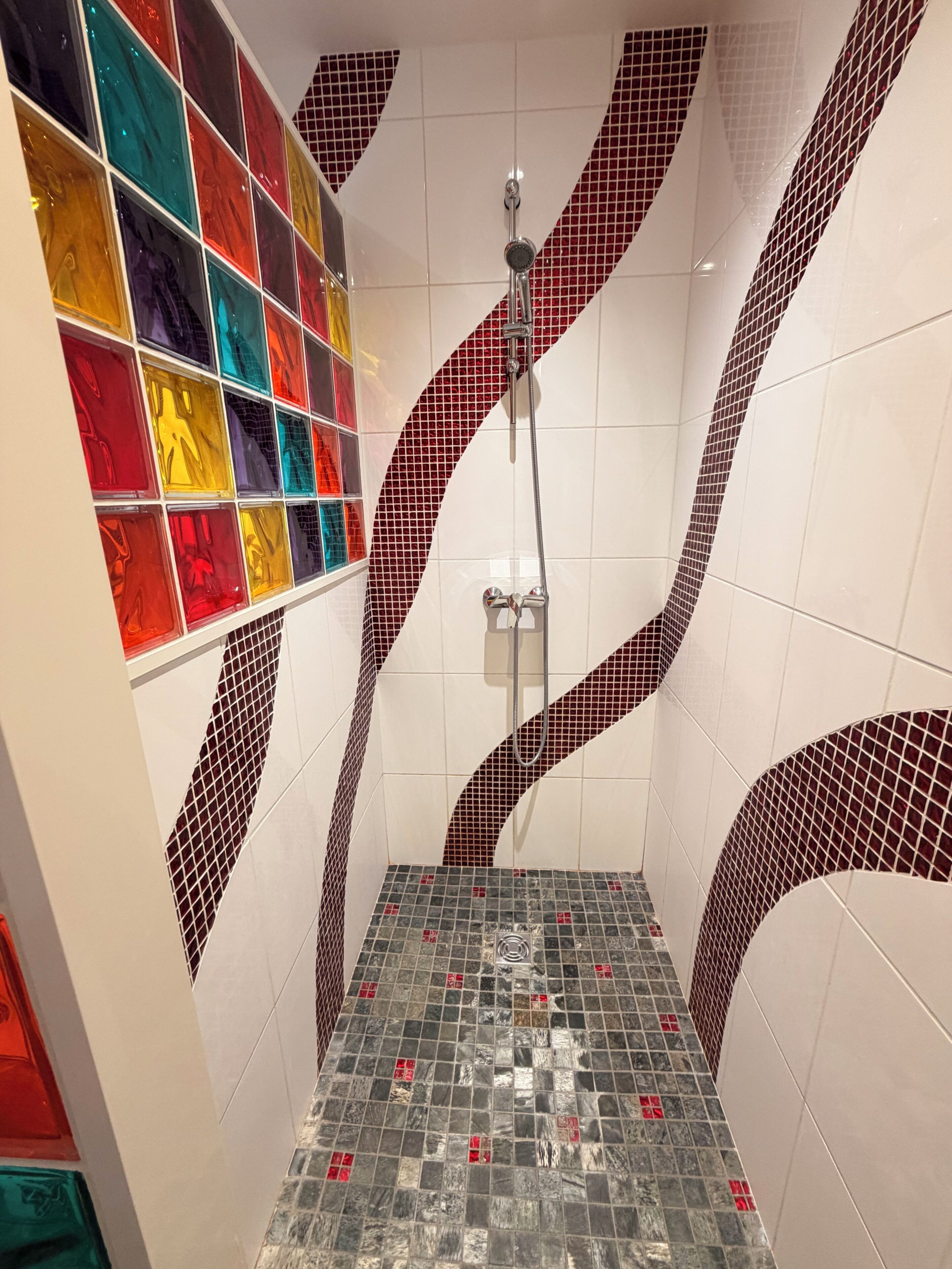 Eine Dusche mit einem einzigartigen Mosaikfliesen-Design, einer Glasduschkabine und einem gefliesten Boden.