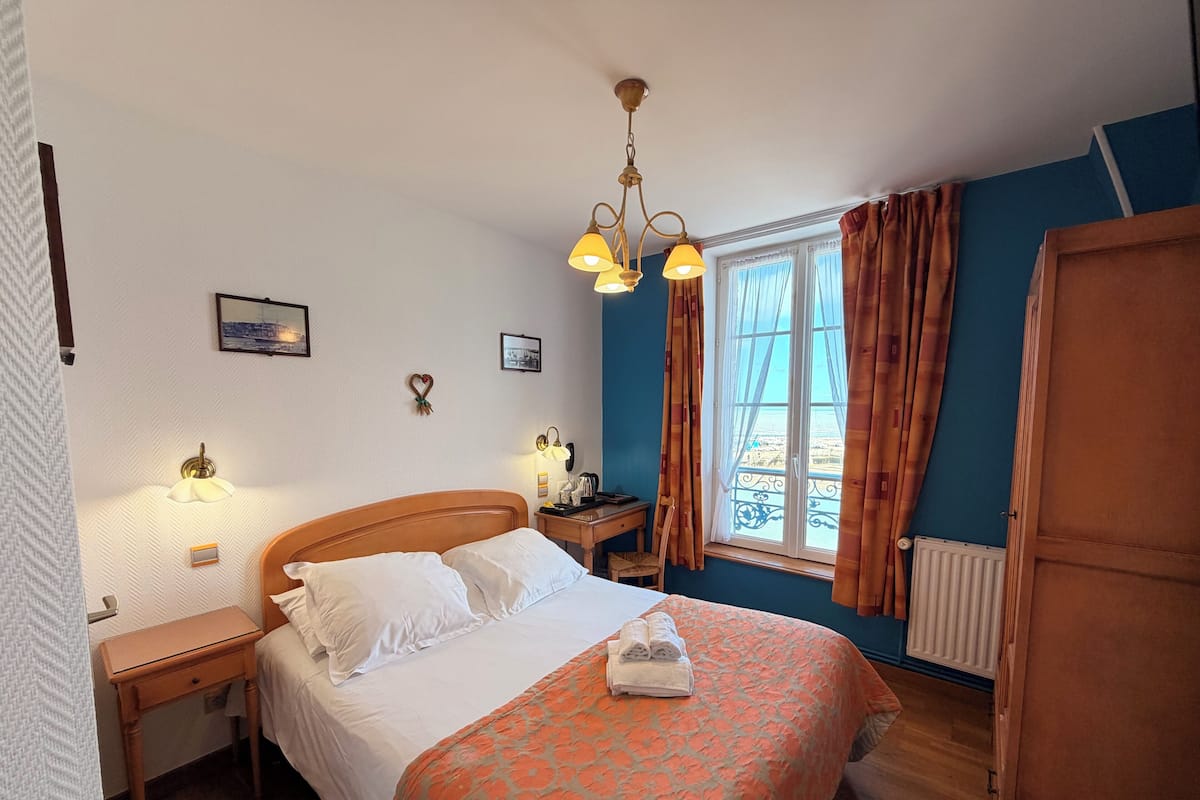 A bedroom with a bed, bedside tables, a nightstand, a window with curtains, a radiator, and a chandelier.