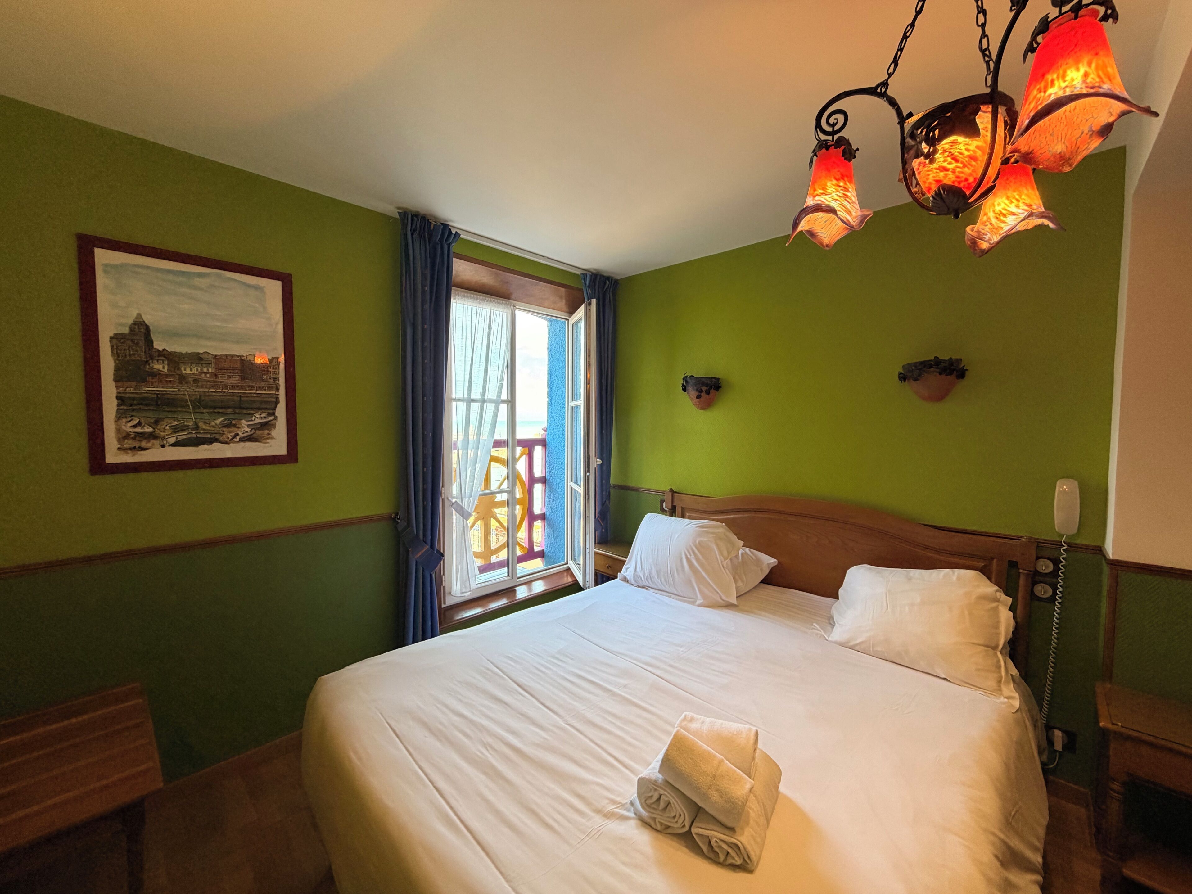 A hotel room with a bed, a window with curtains, a painting on the wall, and a telephone.