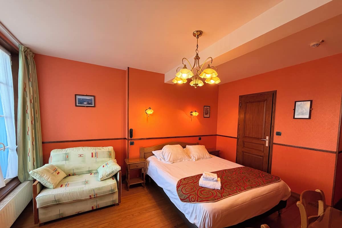 A hotel room with a bed, a sofa, a bedside table, a lamp, and a door.