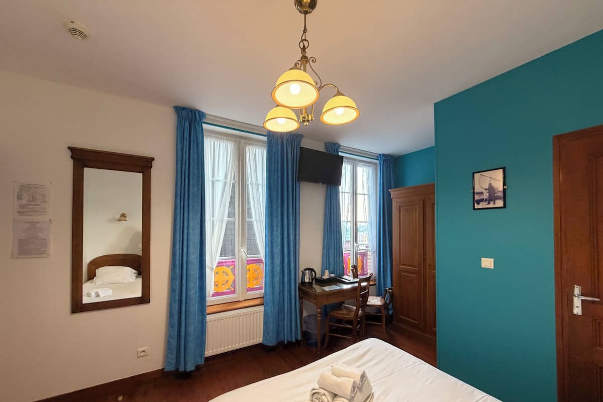 A hotel room with a bed, a desk, a chair, a window with curtains, a radiator, a ceiling light fixture, a door, and a wall-mounted picture.