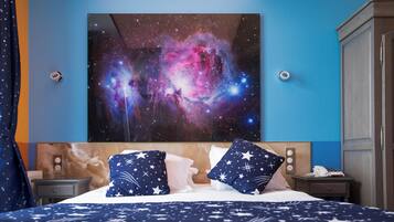 A hotel room with a large galaxy-themed painting on the wall, a bed with star-patterned pillows, and a nightstand with a phone and a lamp.