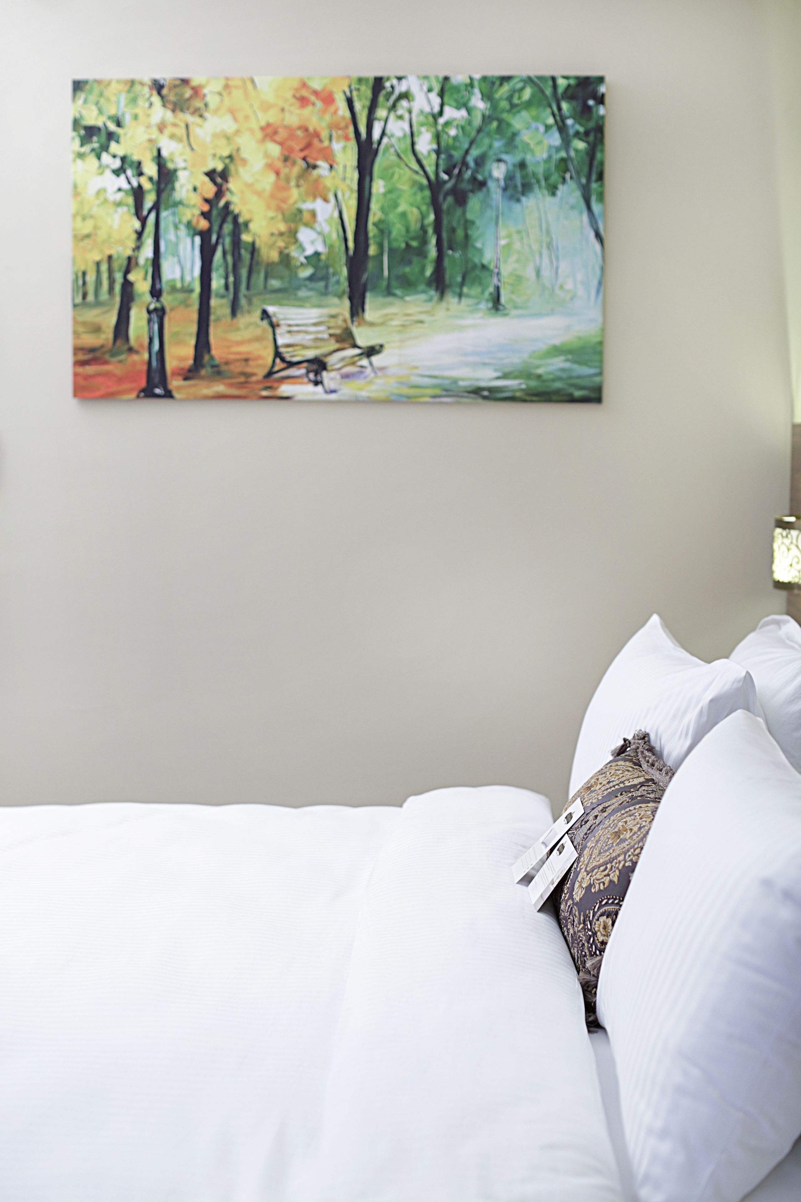 A bedroom with a bed, pillows, and a painting of a park scene on the wall.