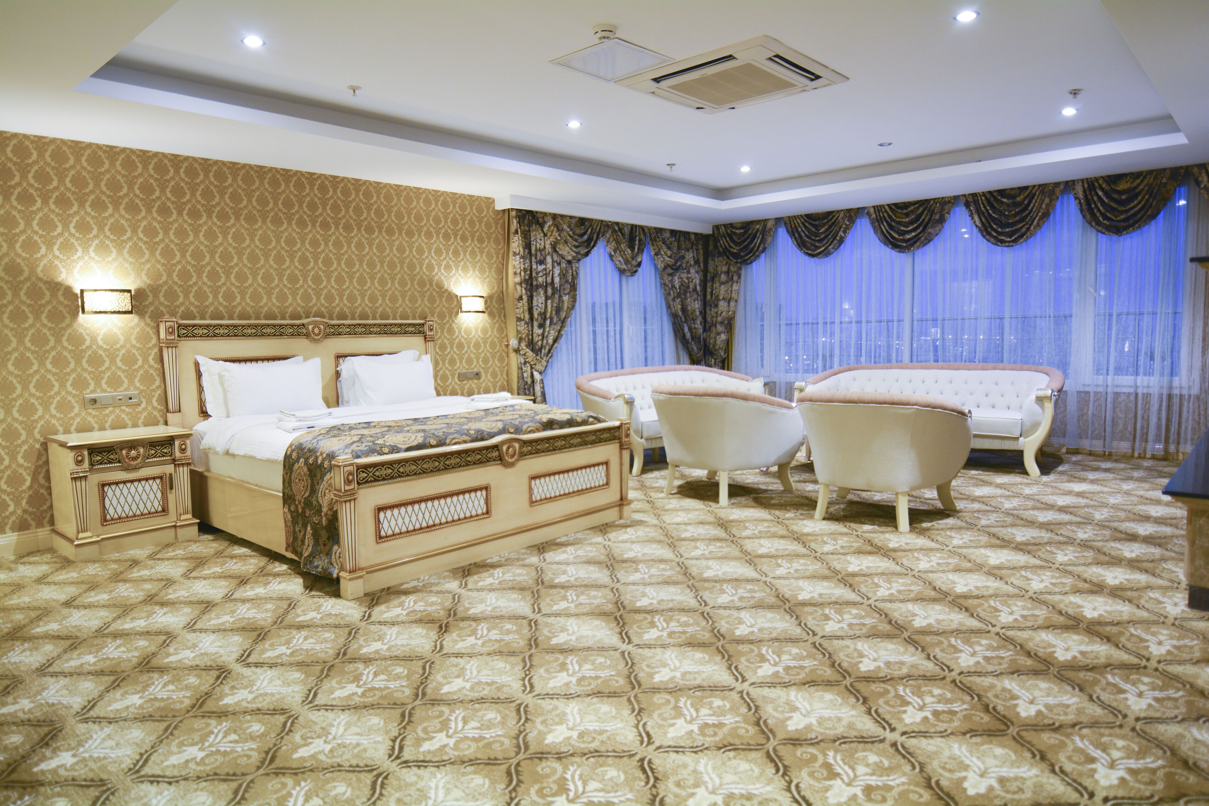 A spacious bedroom with a large bed, two chairs, a vanity, and a large window with curtains.