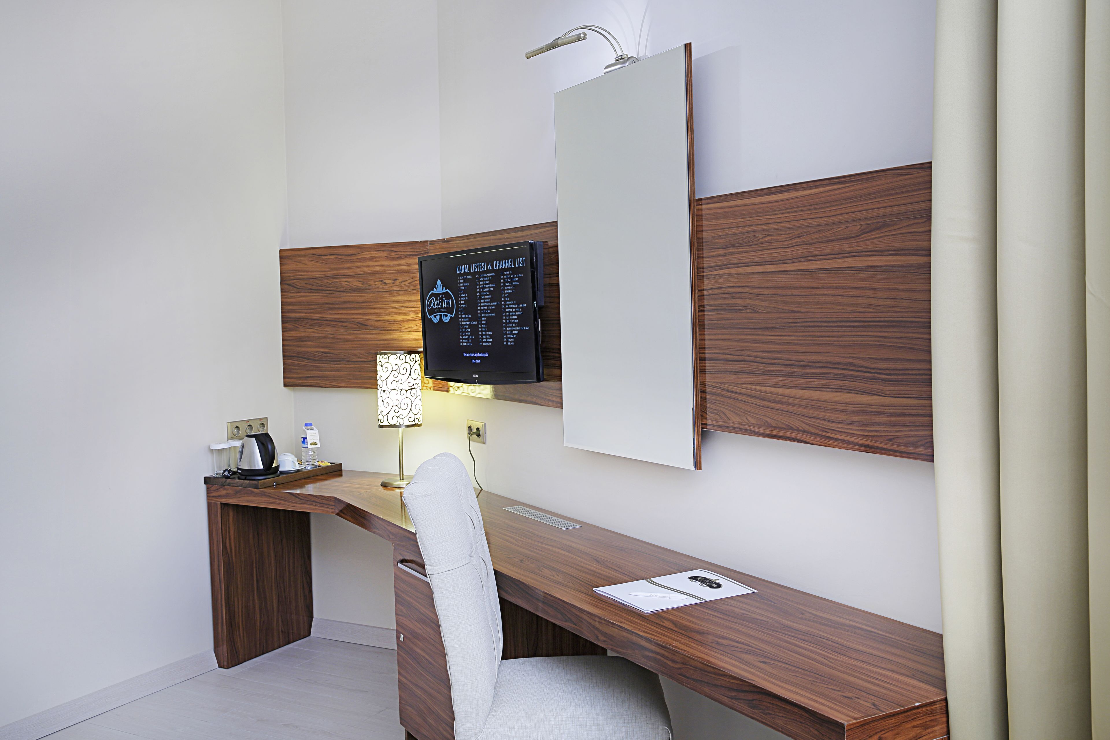 Una habitación de hotel moderna con un escritorio de madera, una silla blanca, un televisor de pantalla plana montado en la pared y una mesita de noche con lámpara.