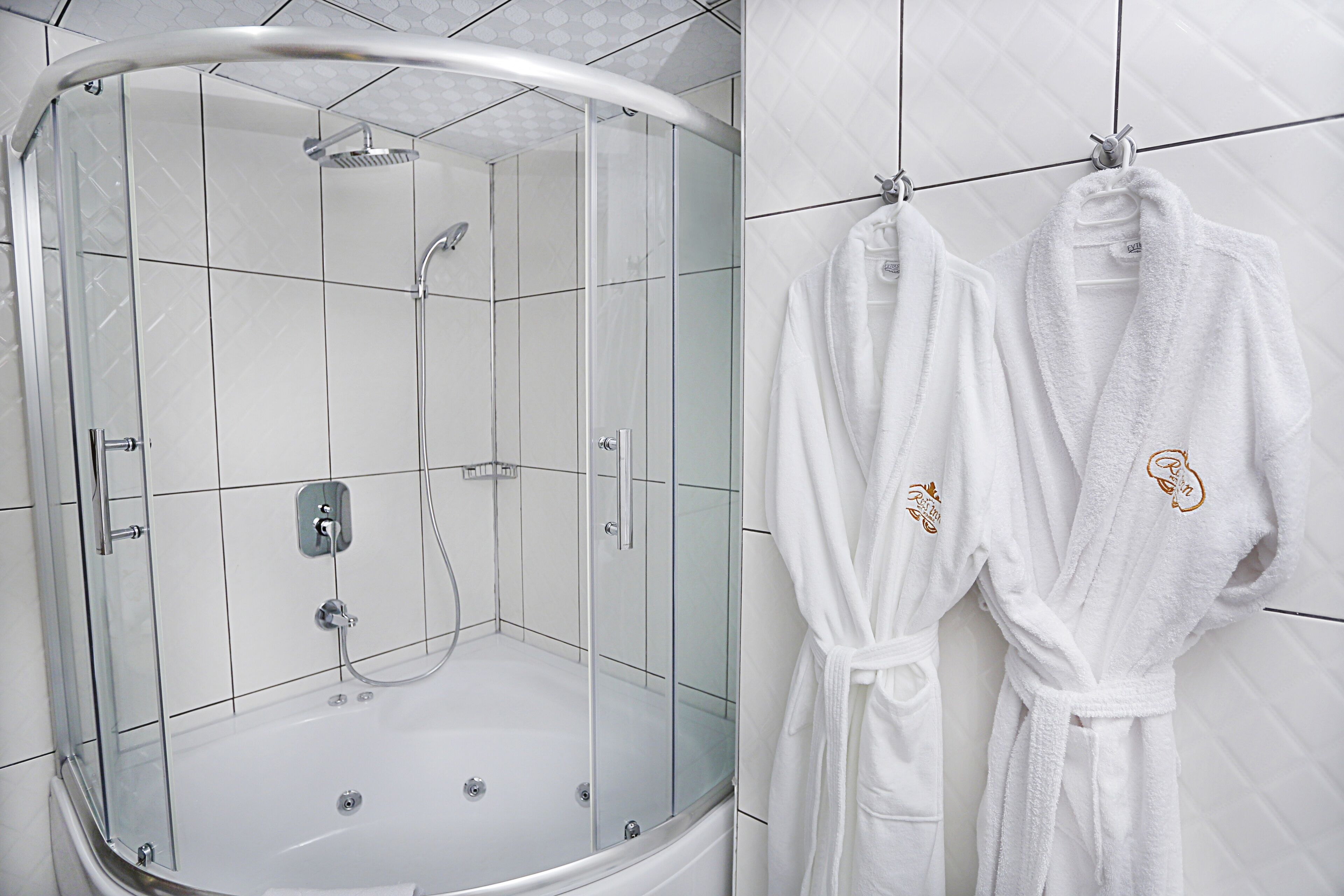A modern bathroom with a glass-enclosed shower, white tiled walls, and two white bathrobes hanging on hooks.