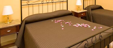 Una cama doble con pétalos de rosa sobre el edredón, una mesita de noche con lámpara y un cuadro en la pared.