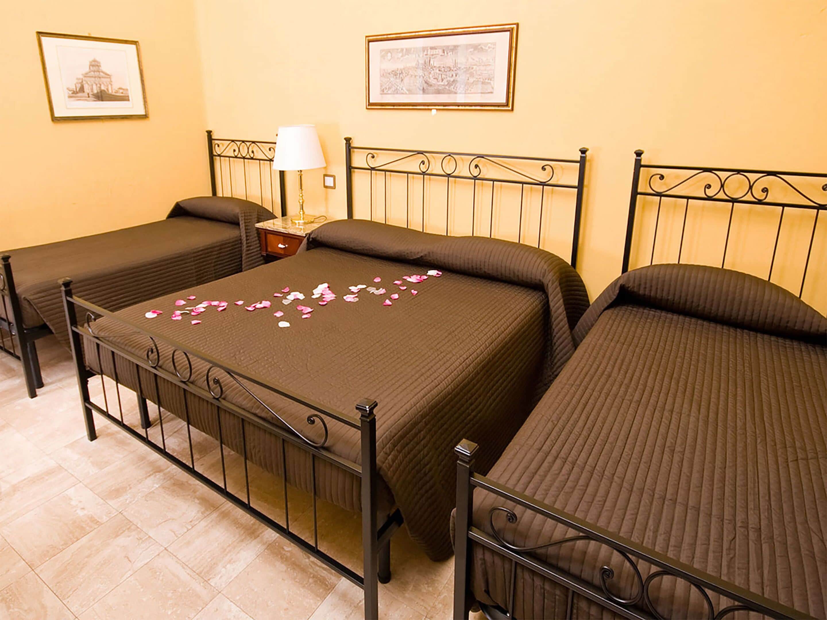 A hotel room with three single beds, a nightstand with a lamp, and two framed pictures on the wall.