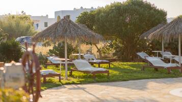 A sunlit outdoor lounge area with thatched umbrellas, stone flooring, and reclining lounge chairs.