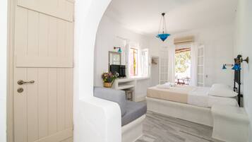 A room with a white door, a grey sofa, a bed with white bedding, a small table with a basket of flowers, and a hanging blue light fixture.