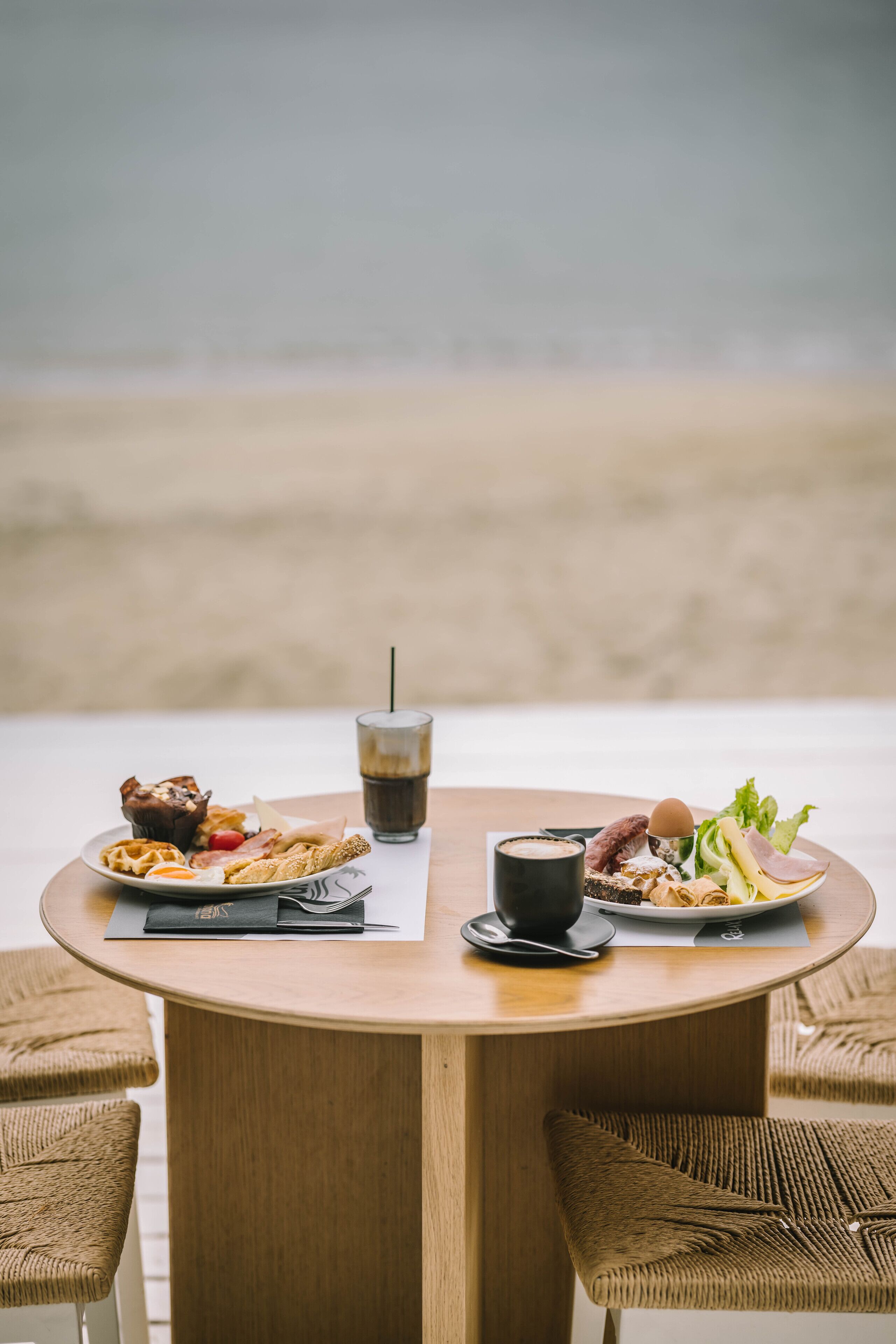 Una mesa redonda de madera con dos platos de comida y una bebida, rodeada de sillas de mimbre.