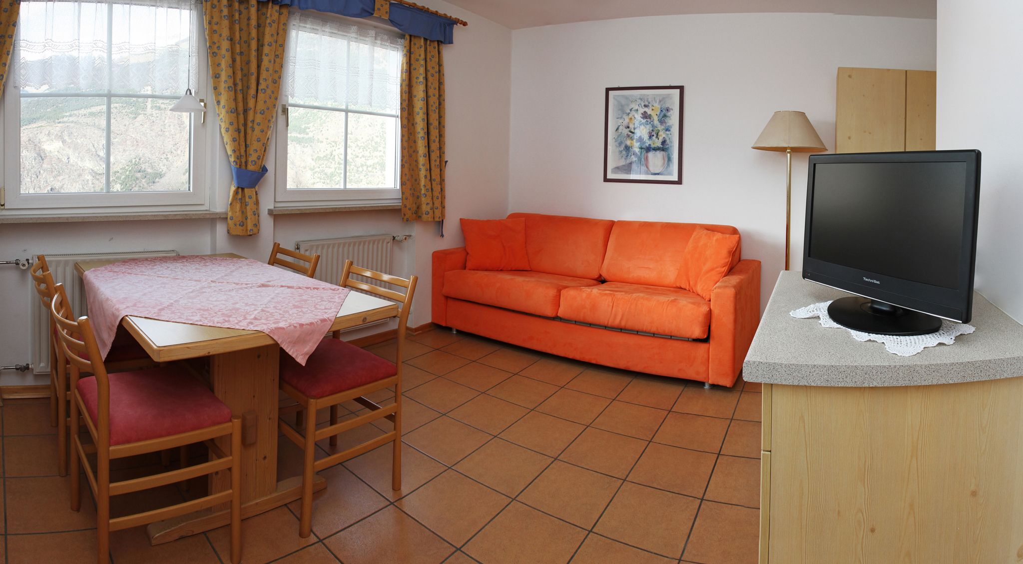 A compact living space with a dining table, orange sofa, and a TV on a wooden stand.